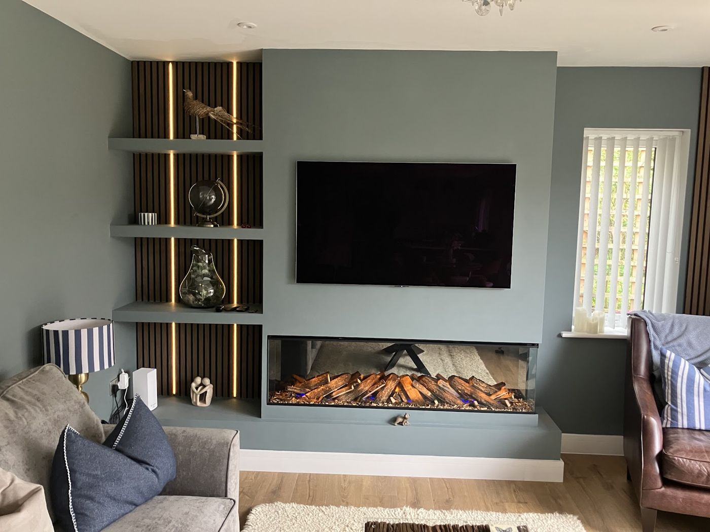 Fitted media wall with a wide inset log-effect fire, wall mounted television and a lit slatted oak panel with floating shelves